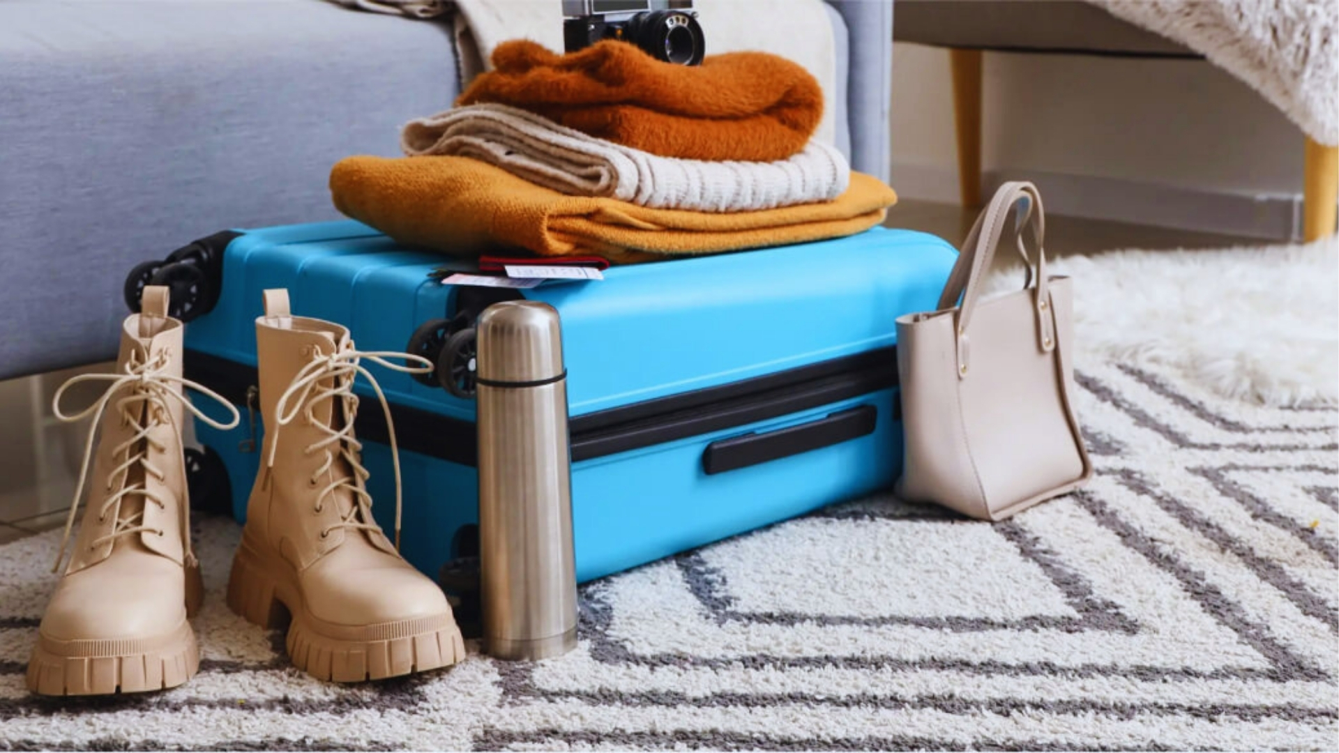 Bright blue hard-shell suitcase packed with wool sweaters, beige combat boots, thermos, and handbag for layering in Iceland weather