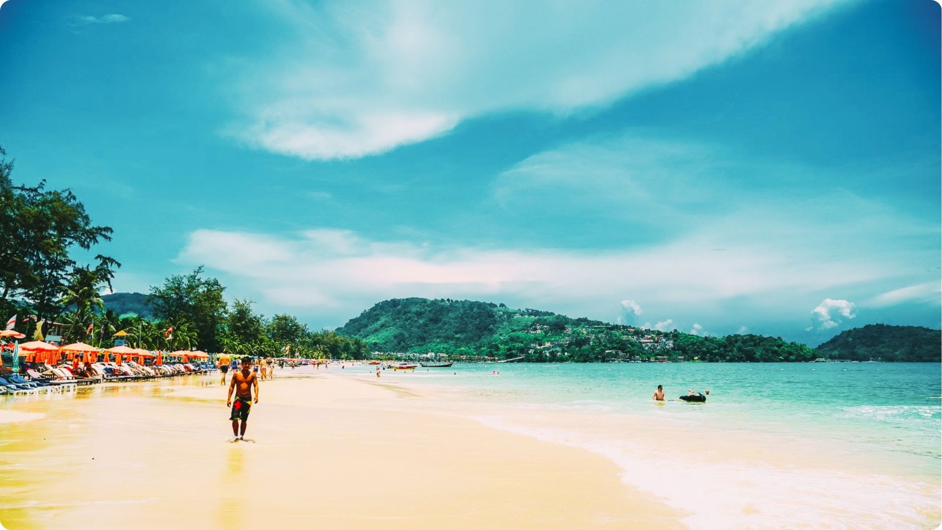 Tourists stroll along a wide sandy beach lined with orange umbrellas and sunbeds.