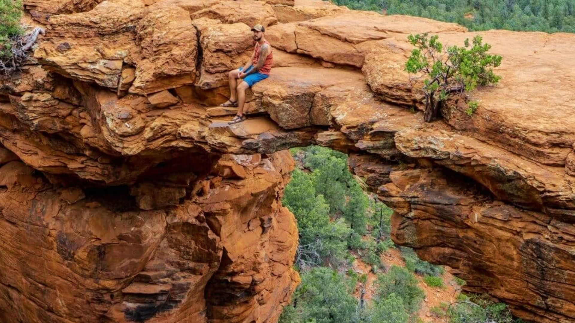 Man sitting on edge of Devils Bridge arch overlooking green forest valley in Sedona Arizona