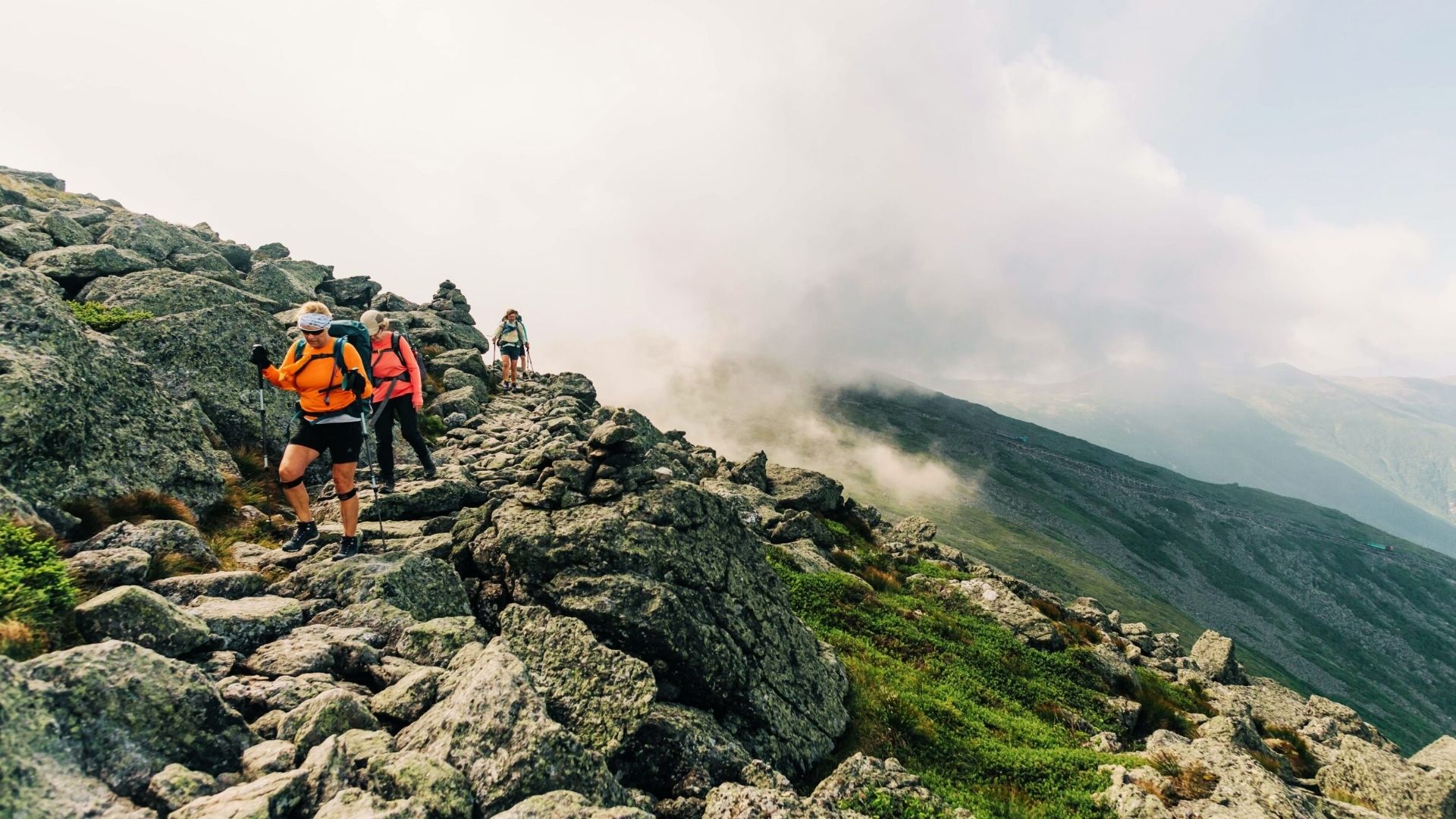 Hikers with trekking poles cross rocky alpine ridge above Ice Lake Colorado trail with misty mountain views and clouds.