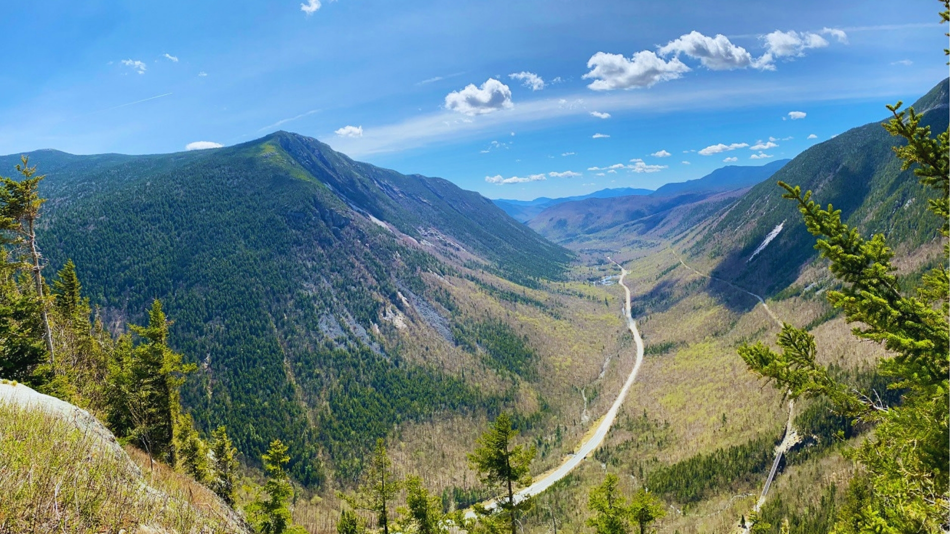 Sunny spring view from Mount Willard Trail shows Crawford Notch with highway, railroad, and forested slopes under blue sky.