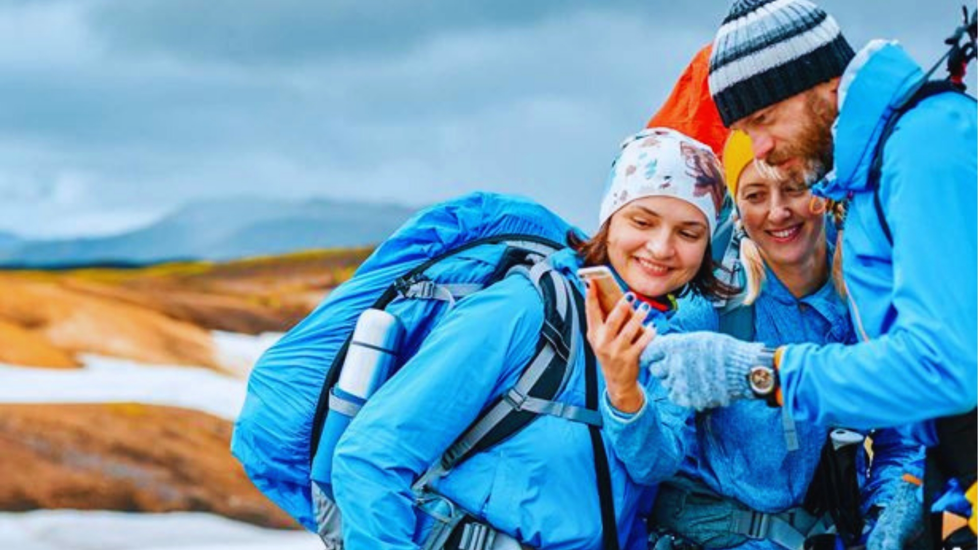 Three hikers in blue waterproof jackets and beanies check phone navigation on snowy Icelandic trails with mountains behind them.
