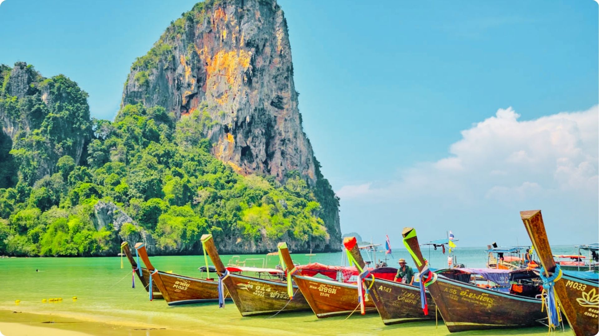 Colorful longtail boats rest on golden sand with massive limestone cliffs rising behind them.