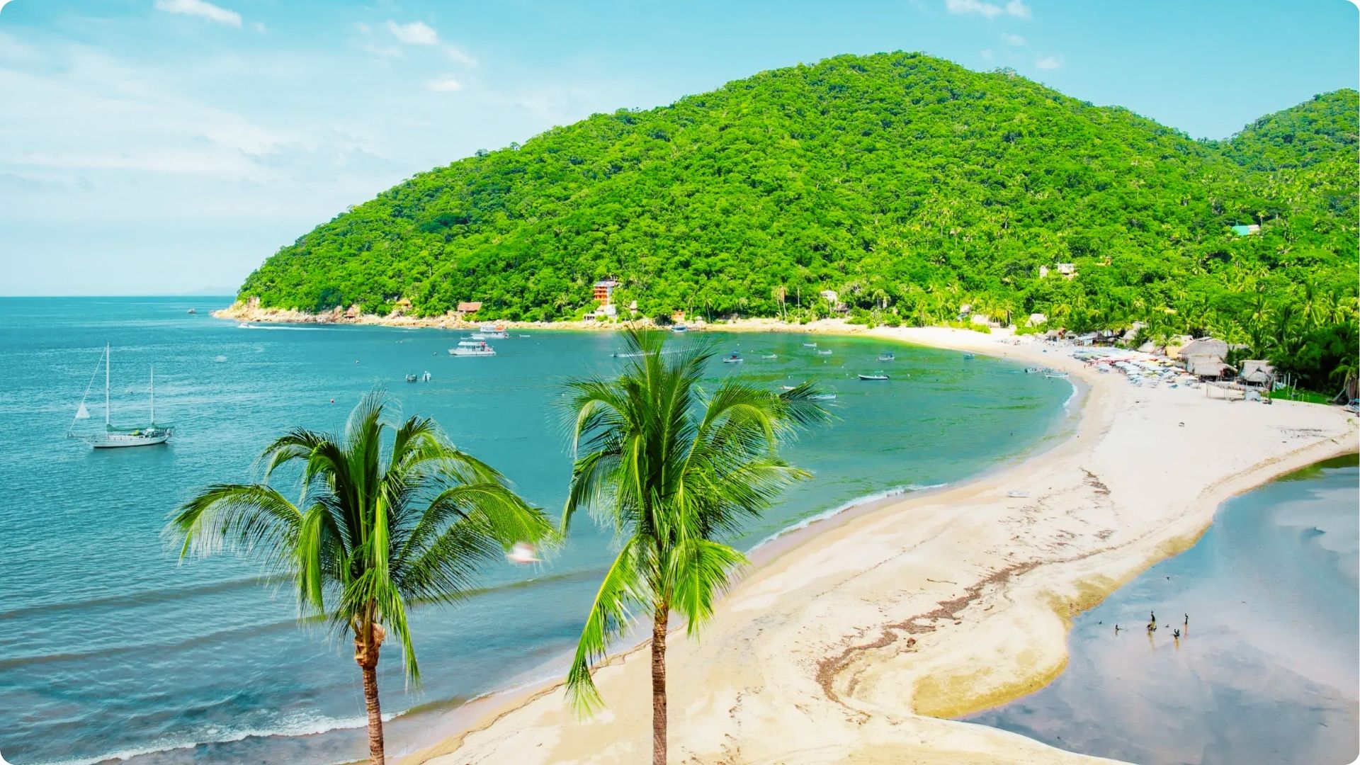 A curved golden sand beach wraps around a calm bay with boats anchored in clear blue-green water.