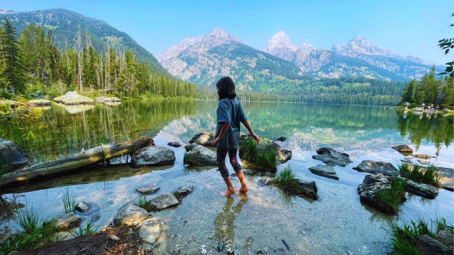 Child wades barefoot through shallow crystal-clear water at Taggart Lake with Teton peaks reflecting on calm surface