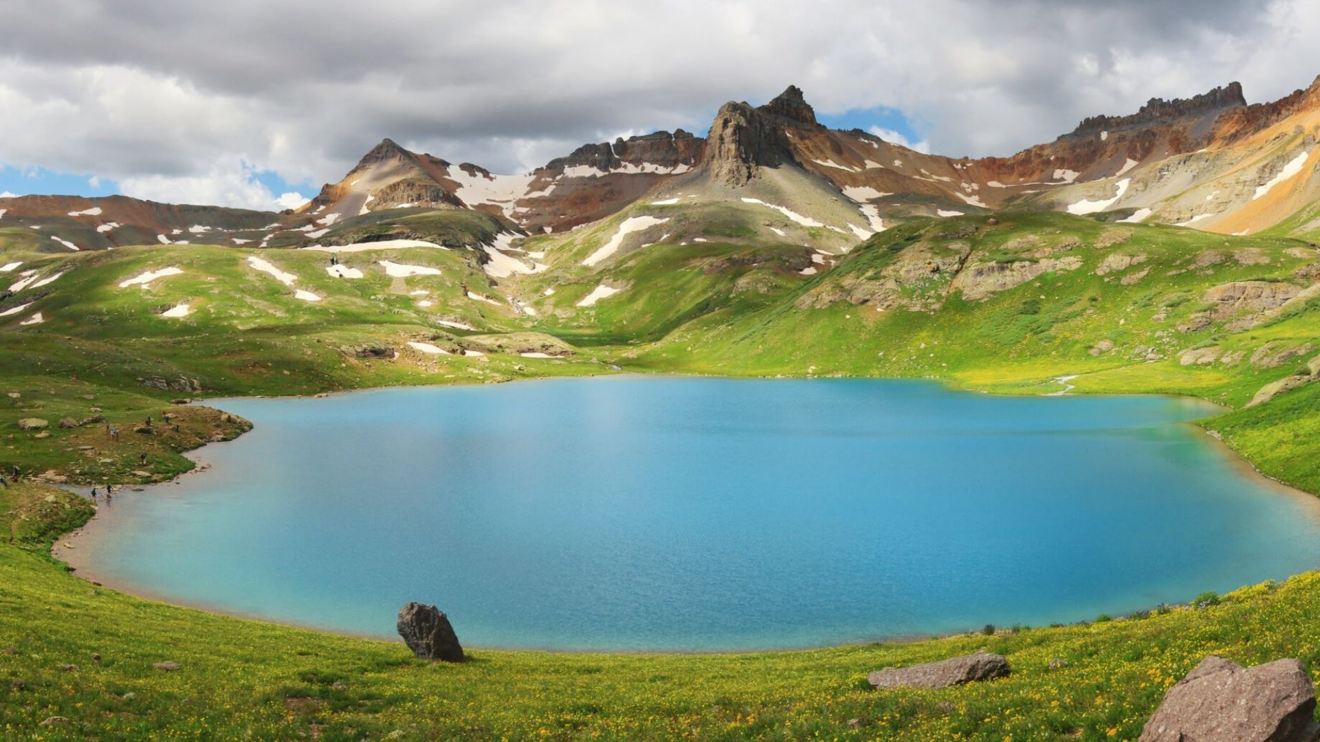 A wide view of Ice Lake Colorado’s vivid blue water surrounded by green slopes, yellow wildflowers, and dramatic mountain ridges.