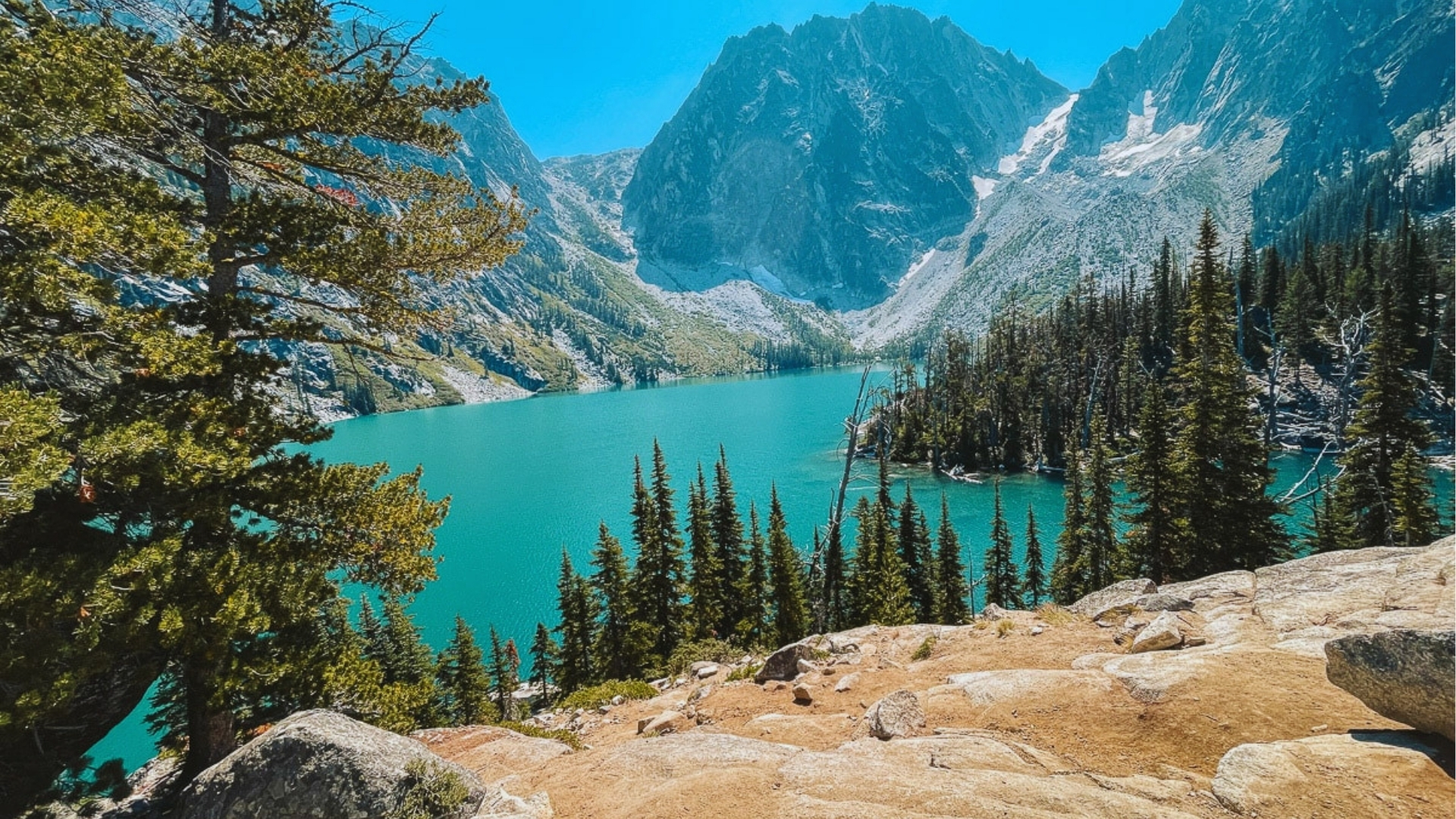Bright summer scene of Colchuck Lake from granite overlook with vivid turquoise water, pine trees, and Dragontail Peak looming.