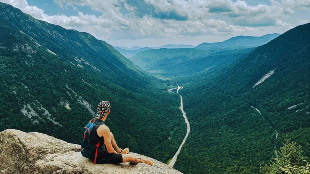 Hiker with patterned hat sits on granite ledge overlooking lush green Crawford Notch valley and winding road from Mount Willard.