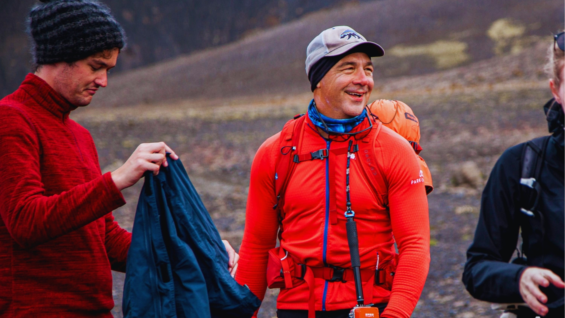 Group of hikers in red thermal layers and backpacks adjust gear on volcanic Iceland terrain wearing beanies and windproof jackets