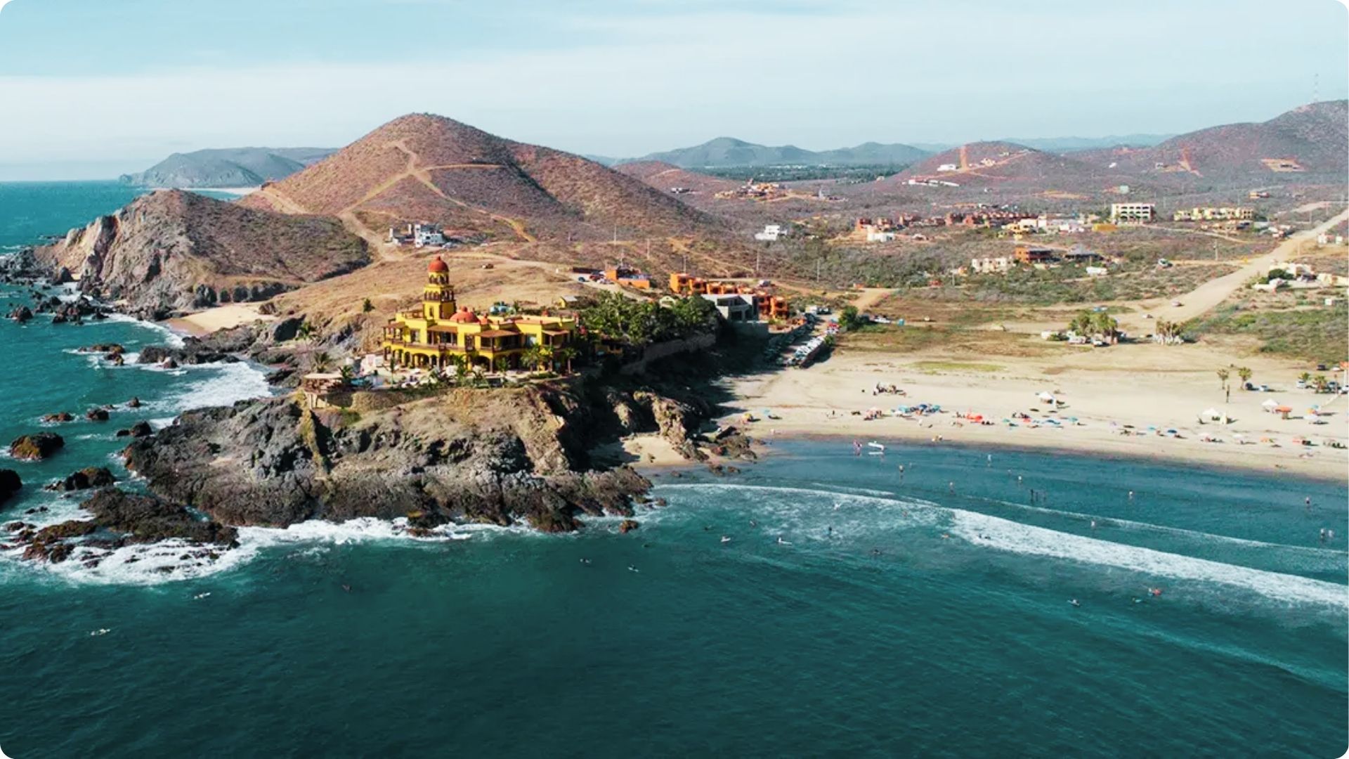 A yellow colonial-style villa sits atop a rocky cliff overlooking a wide sandy surf beach.