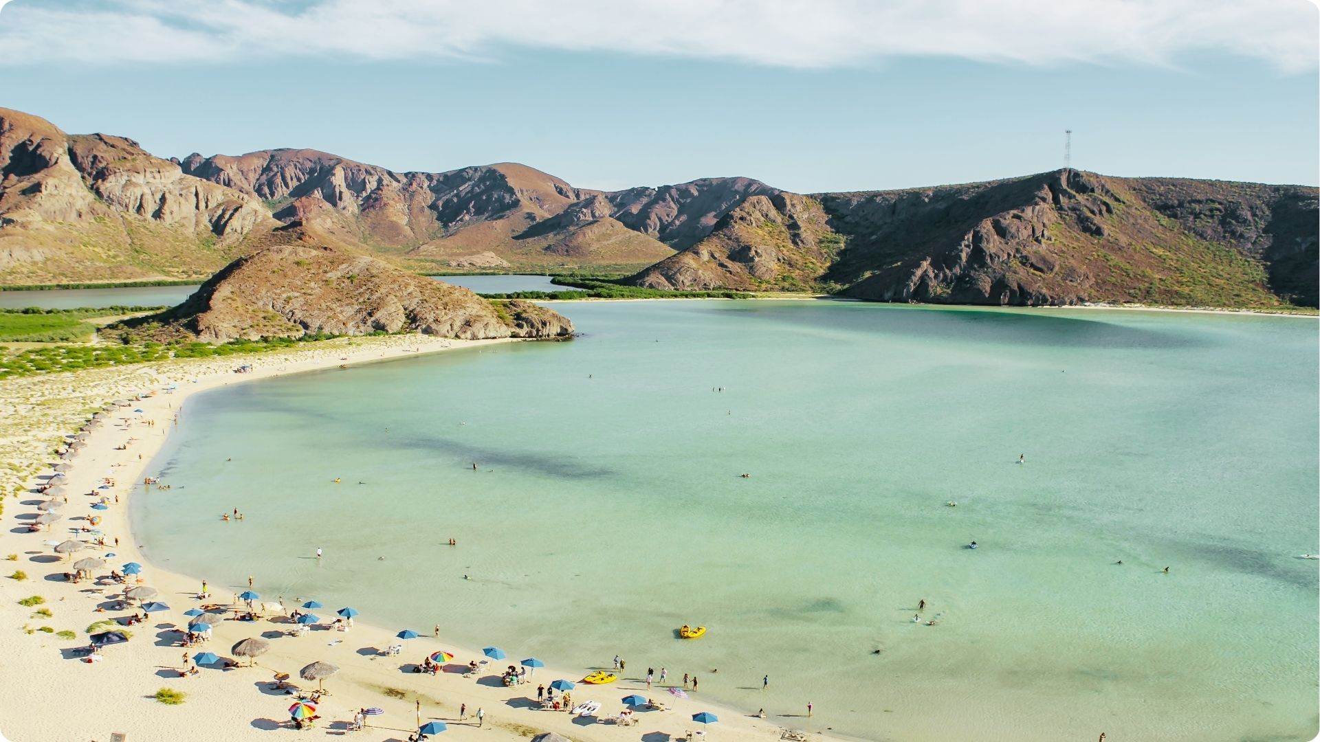 A crescent-shaped bay with shallow, clear aqua water is surrounded by red desert mountains.