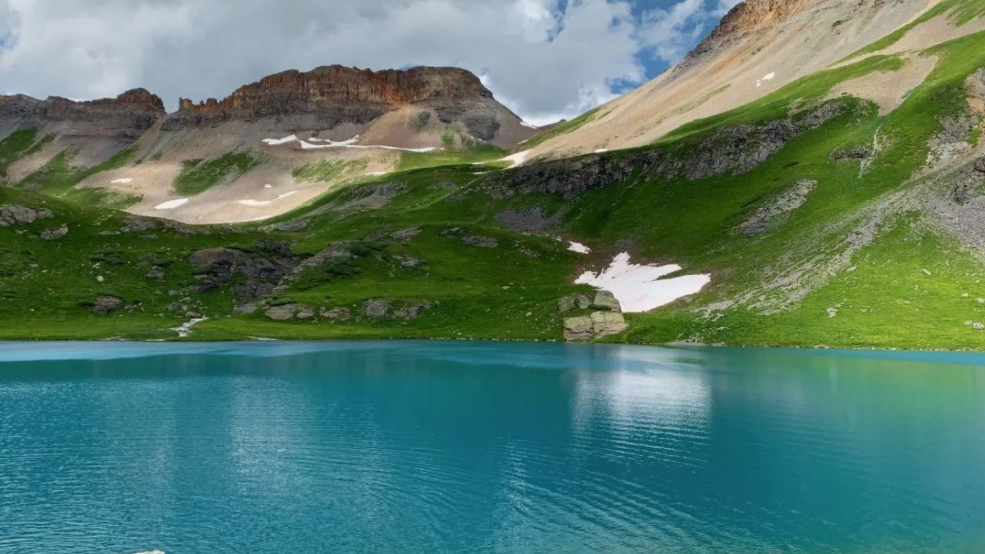 Panoramic view of bright turquoise Ice Lake Colorado ringed by green meadows, snow patches, and jagged San Juan peaks.
