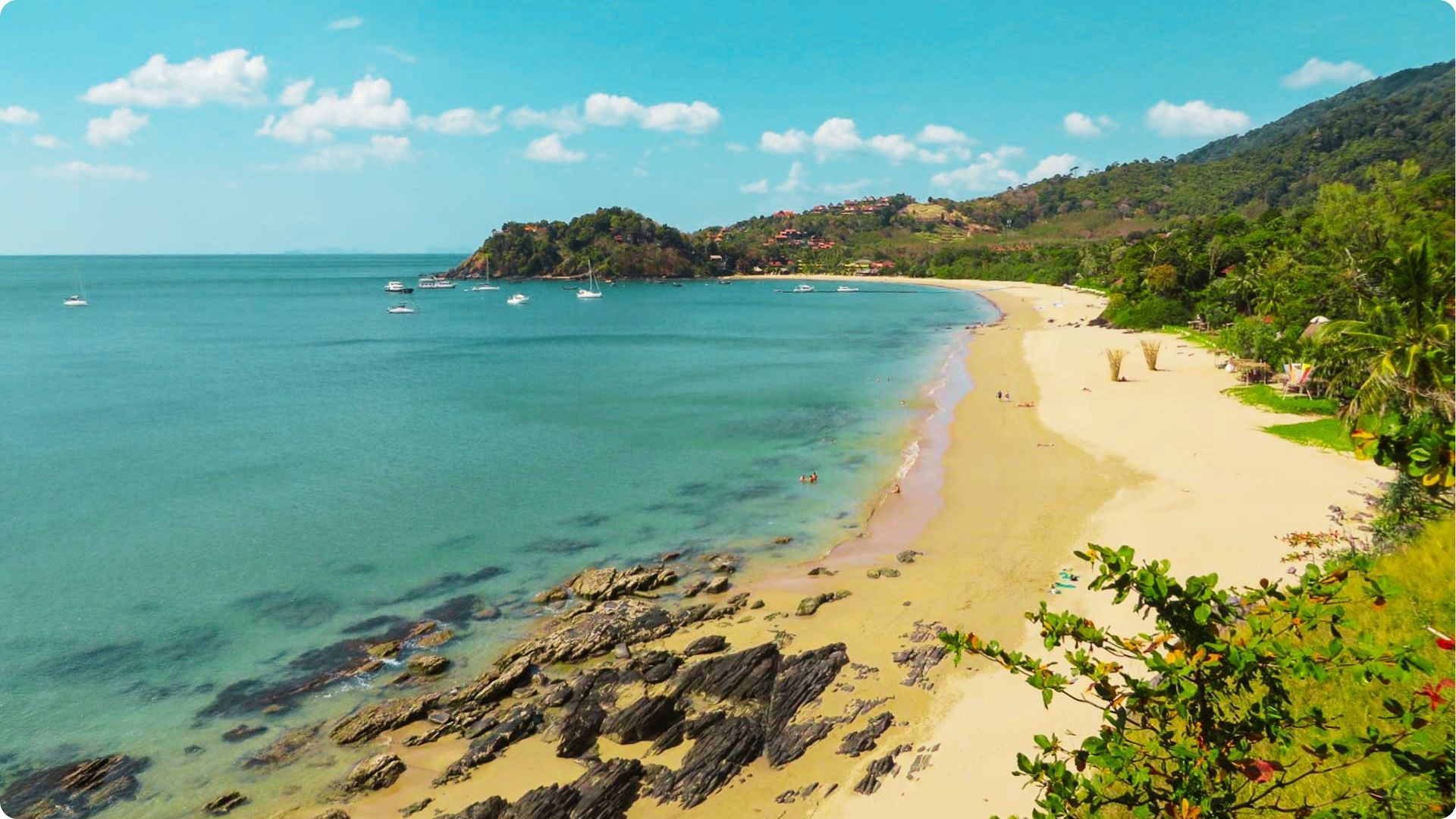 A quiet, crescent-shaped beach with golden sand meets clear aqua water under a bright blue sky.