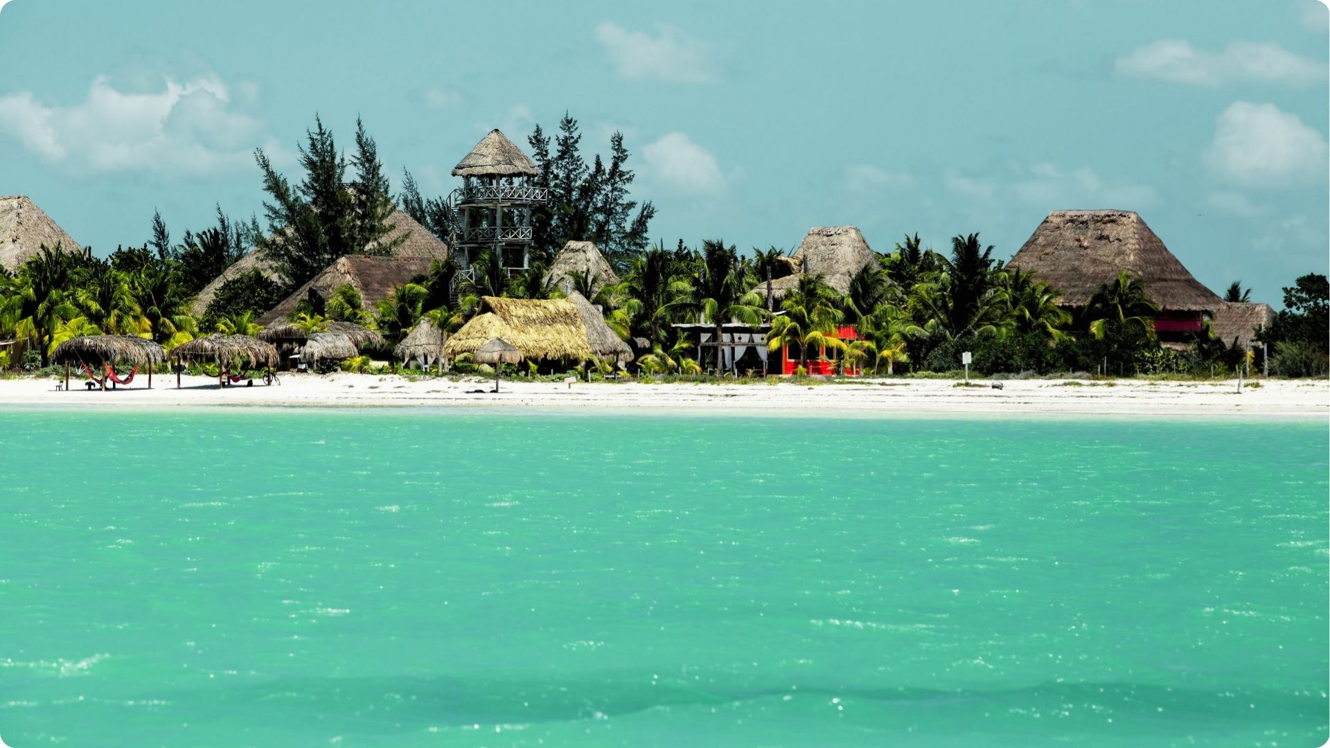 Rustic palapa huts and a lookout tower sit on a white sand beach facing bright turquoise water
