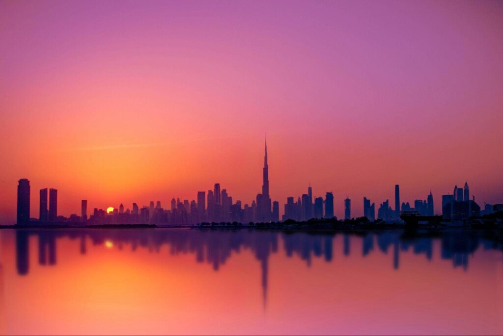 “UAE on a Budget: A Family-Style Plan” . The sun sets behind Dubai's skyline, casting a warm glow over the iconic buildings and reflecting on the water.